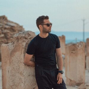 Fashionable man in sunglasses leaning on ancient stone ruins with a serene background.